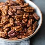 Golden-brown air fryer candied pecans in a bowl with cinnamon sticks beside them.