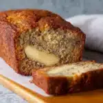 A golden-brown loaf of cream cheese banana bread sliced on a wooden cutting board.