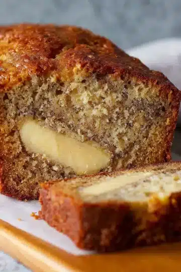 A golden-brown loaf of cream cheese banana bread sliced on a wooden cutting board.