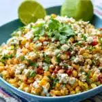 A bowl of creamy Mexican Corn Salad garnished with cotija cheese, cilantro, and lime wedges.